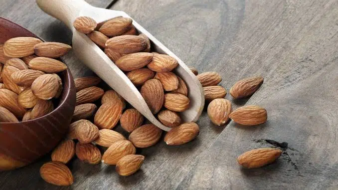 Amandes disposées dans une petite pelle en bois clair et dans un bol en bois foncé. Quelques amandes sont posées directement sur le plan de travail en bois.