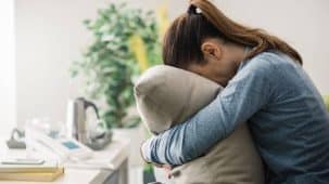 Une jeune femme anxieuse et stressée serre un coussin dans ses bras, la tête baissée. Elle est assise sur son canapé, porte une queue de cheval et un pull bleu.