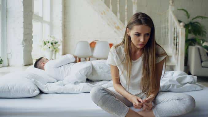 Anxiolytiques naturels : une jeune femme est anxieuse au bord du lit. On aperçoit son compagnon dormir derrière elle.