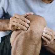 Un homme assis souffre d'arthrose à son genou qu'il tient entre ses deux mains. Il porte un short noir et un t-shirt gris.