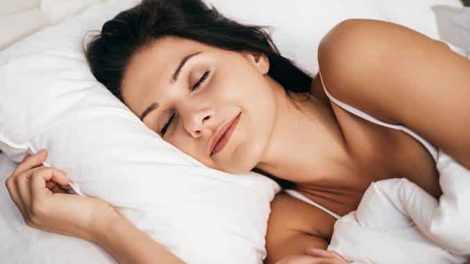 Femme en train de dormir paisiblement, le sourire aux lèvres, sur un lit, oreiller, couette blancs.