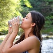 Une femme brune boit de l'eau pure dans un verre entourée de nature et d'une rivière