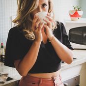 Une femme blonde bois une infusion minceur dans sa cuisine. Elle porte un t-shirt noir au dessus du nombril et un pantalon couleur brique.