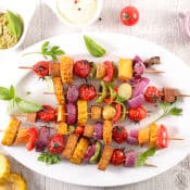 Brochettes végétales (tomate, maïs, tofu, oignon, poivron, pomme de terre) posées sur une assiette ovale blanche. Fond en bois clair.