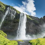 Cascade dans un endroit naturel. L'eau jaillit d'un rocher et tombe dans un plan d'eau entouré de nature verdoyante. Le ciel est bleu.