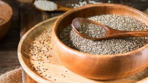 Graines de chia dans une cuillère et un bol en bois. L'ensemble est posé sur une assiette en bois posée sur un plan de travail en bois.