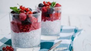 Pudding de chia dans deux verres disposés sur une table en bois et sur un torchon à carreaux bleus et blancs. Le mélange chia et lait végétal est recouvert d'un coulis de fruits rouges, de framboises, de mûres, de groseilles et de menthe.