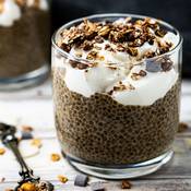 Une verrine remplie de pudding de chia au chocolat, avec une couche de fromage blanc saupoudrée de céréales croustillantes. La verrine est posée sur une table en bois, il y a une cuillère en métal à côté de la verrine.