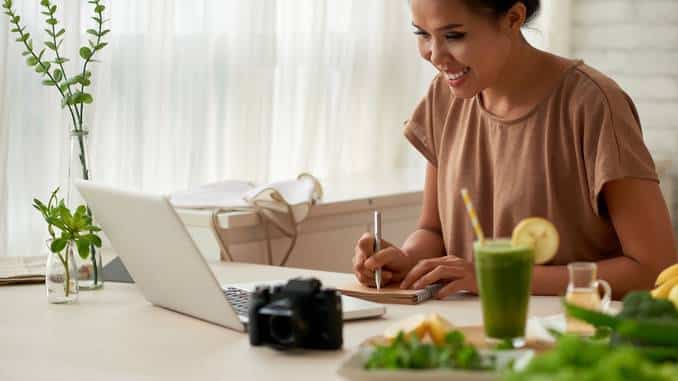 Une femme brune qui écrit sur du papier devant son ordinateur en souriant. Il y a un smoothie vert, des plantes et un appareil photo sur la table