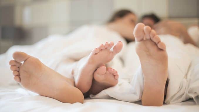 Couple homme / femme allongé sur un lit, sous une couette blanche. Gros plan sur leurs pieds qui se câlinent.