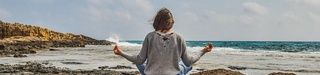 Femme brune assise en tailleur face à la mer, sur une plage. Elle semble méditer, avec les paumes de ses mains vers le ciel, cheveux au vent.