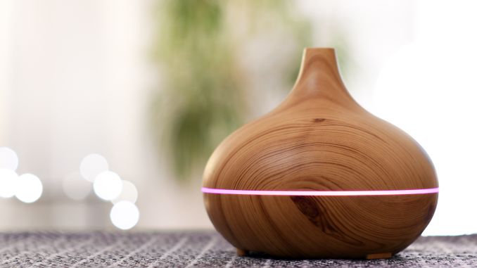 Diffuseur d'huiles essentielles en gros plan. Fond flou avec plante. Il s'agit d'un brumisateur en bois avec une ligne horizontale de lumière rose en son centre. Un filet de brume s'échappe de l'appareil.