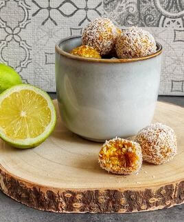 Energy balls végétales et sans gluten posées dans une tasse sur une rondelle de bois. Les boules sont jaunes et enrobées de noix de coco.