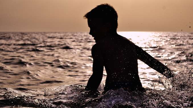 Enfant dans la mer : le garçon apparaît en contre-jour avec le reflet du soleil dans l'eau derrière lui.