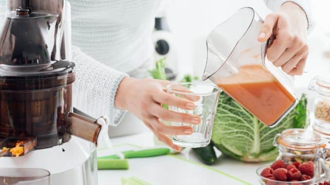 Gros plan sur les mains d'une femme sur le point de verser un jus frais de couleur orange dans un verre. Un extracteur de jus vertical, une planche à découper, ainsi que plusieurs fruits et légumes sont posés sur le plan de travail blanc.