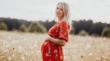 Femme enceinte dans la nature. Elle porte une robe rouge, se tient de profil et pose sa main sur son ventre. Elle se trouve dans un champ avec une forêt au loin.