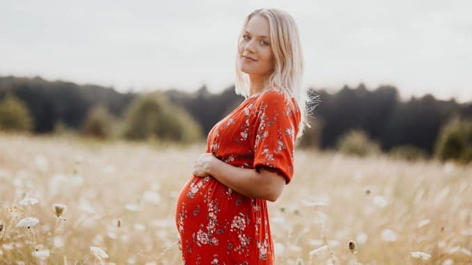 Femme enceinte dans la nature. Elle porte une robe rouge, se tient de profil et pose sa main sur son ventre. Elle se trouve dans un champ avec une forêt au loin.