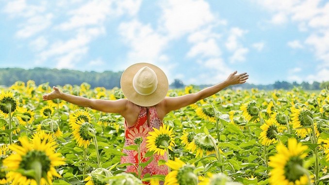 Une femme de dos, les bras en l'air, qui porte un chapeau de paille en plein milieu d'un champ de tournesols en fleur. Le ciel est bleu avec quelques nuages.