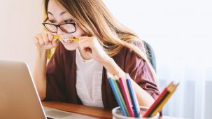 Une femme assise à son bureau devant son ordinateur avec un stylo dans la bouche, stressée et crispée. Rideau blanc en fond et pot de crayons/stylos en premier plan.