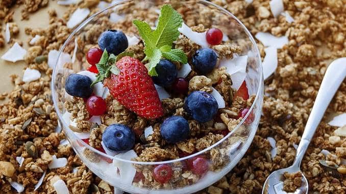 Aliments riches en fibres : il y a un bol en verre rempli de muesli sur une table recouverte de céréales. Le bol de muesli contient également des myrtilles, des fraises ainsi que des copeaux de noix de coco et une feuille de menthe fraîche.