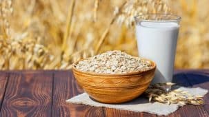 Flocons d'avoine dans un bol en bois et verre de lait d'avoine posé sur une table en bois. En arrière plan, on aperçoit un champ d'avoine.