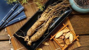 Racines de ginseng dans un récipient noir, présentés sur une table en bois. Des tranches de ginseng sont posées à côté.