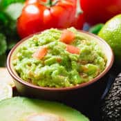 Guacamole dans un ramequin. On distingue clairement la purée d'avocats et les tomates. Autour du ramequin il y a des avocats, citrons, tomates, feuilles de coriandre...