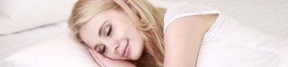 Femme blonde qui dort sur le côté, le sourire aux lèvres, habillée en blanc. Elle pose sa tête sur ses mains, elles-mêmes posées sur son oreiller blanc.