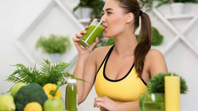 Gros plan sur une femme qui boit un jus vert dans un verre. Tout autour, il y a des fruits et légumes : pommes vertes, citrons, poivron jaune, brocolis.