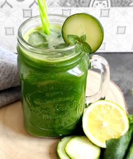 Jus vert servi dans un verre posé sur une rondelle de bois. Il y a un concombre placé sur le dessus du vert et une feuille de menthe.