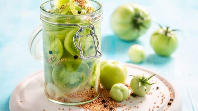 Lacto-fermentation : bocal de tomates vertes lactofermentées sur une table en bois. On aperçoit des graines de moutarde et des baies de poivre noir sur la table.