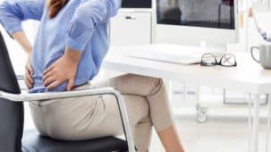 Femme qui a mal au dos assise sur une chaise face à un bureau. Elle pose ses mains sur le bas de son dos.
