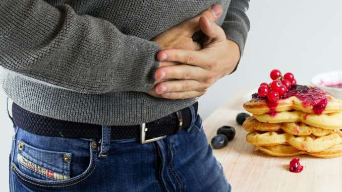 Homme qui se tient le ventre avec les deux mains sur son ventre. Plan de près, on aperçoit seulement le haut de son jean, sa ceinture et son ventre sous un pull. On imagine une douleur abdominale. Des pancakes sont présentés en arrière plan.