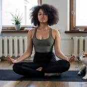 Insomnie et méditation : une femme métisse fait de la méditation dans son appartement ur un tapis au sol. Elle a les jambes qui se croisent et la tête haute.