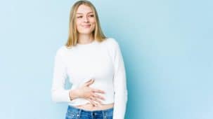 Microbiote et dépression : une jeune femme blonde pose sa main sur sa ventre et sourit. Elle porte un pull blanc et un jeans bleu. On entrevoit son nombril.