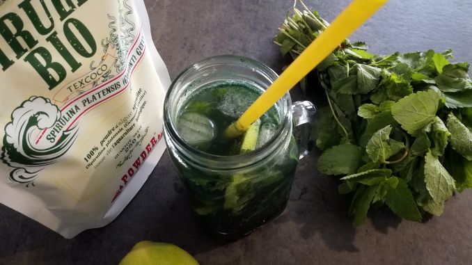 Verre de Mojito Spiruline entouré d'un sachet de spiruline, de feuilles de menthe et d'un citron.