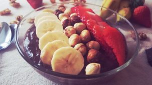 Crème type mousse banane chocolat cacao dans un bol décoré de rondelles de banane, de noisettes et de fraise coupée en tranches.