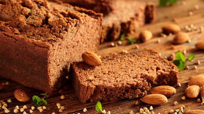 Pain sans gluten avec une tranche coupée posée sur le support en bois. Il y a aussi des amandes.