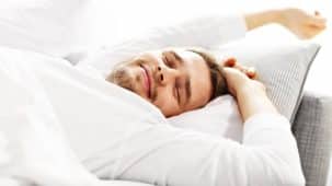 Homme en train de dormir paisiblement, le sourire aux lèvres, sur un lit, oreiller, couette blancs. La luminosité de l'image fait penser au bien-être.