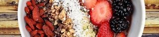 Bol rempli de fruits crus et fruits secs disposés en lignes (amandes, fraises, mûres, coco...). Le bol blanc est posé sur une surface en bois.