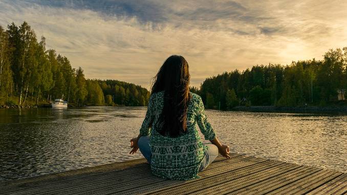 Femme assise en tailleur face à un paysage naturel (lac, forêt, coucher de soleil). Santé, bien-être et détente.