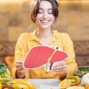 Femme souriante, brune, assise à table, habillée en jaune, tient une représentation de foie dans ses mains. Des aliments sains sont disposés sur la table devant elle. La santé hépatique est mise en avant.