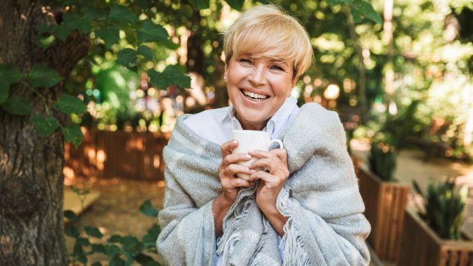 Femme blonde, senior, souriante et heureuse avec sa tasse et son plaid dans un coin de verdure avec arbre et plantes.