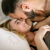 Vitamine B9 et fertilité : un homme (brun avec barbe) et une femme (blonde aux cheveux longs) sont allongés dans un lit. L'homme est positionné au dessus de la femme. Ils se sourient et sont prêts à s'embrasser.