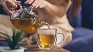 Une femme brune sert du thé dans une tasse en verre posée sur une table en bois. On aperçoit une plante et un canapé bleu.