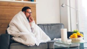 Jeune homme malade qui tousse assis sur un canapé gris, enveloppé dans une couette blanche.