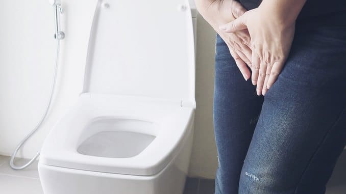 Une jeune femme souffre de vessie hyperactive. Elle a ses deux mains contre son entre-jambe. Elle se trouve à côté des toilettes.