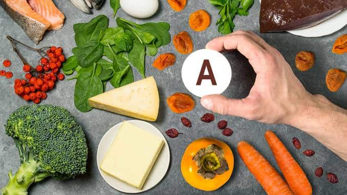 Aliments riches en vitamine A posés sur une table grise : carottes, brocolis, fromage, poissons, kaki, abricots secs, épinards, foie, etc.