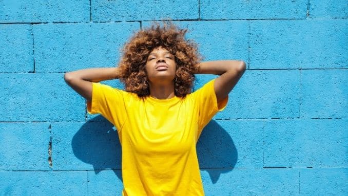 Une femme afro-américaine fait le plein de vitamine D. Elle s'expose au soleil , le dos contre un mur bleu. Elle porte un t-shirt jaune.