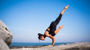 Femme qui pratique le yoga sur un rocher au bord de la mer. Elle soutient son corps sur les mains, à la force de ses bras, jambes en l'air.